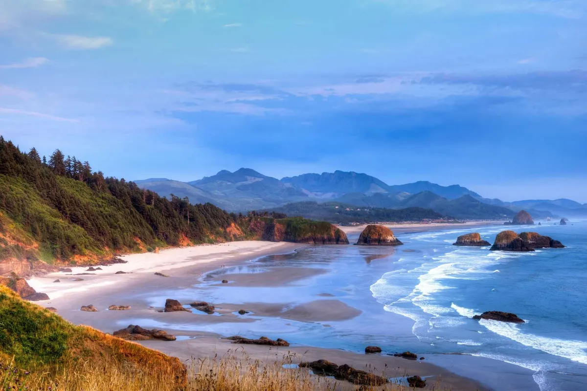 A breathtaking view of unique coastal rock formations, a sandy beach, gentle ocean waves, and a mountainous backdrop. The mood is tranquil and open, perfect for sparking creativity or unwinding after a productive work session.