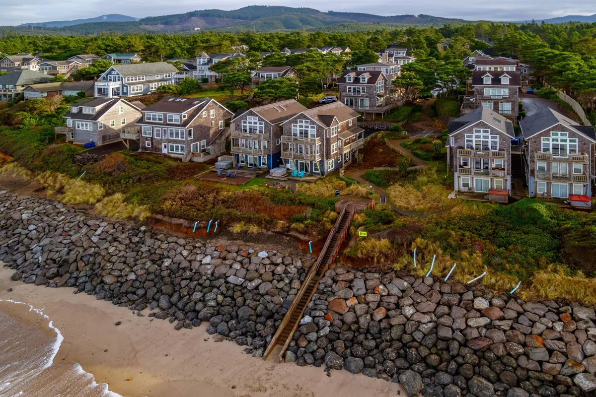 Beachfront cottages line a gently curving, rocky shoreline with a wooden stairway inviting guests directly to the sand. Surfers’ boards and seaside activity highlight the area’s vibe, while the scene suggests bonfires, family picnics, and playful days by the water.