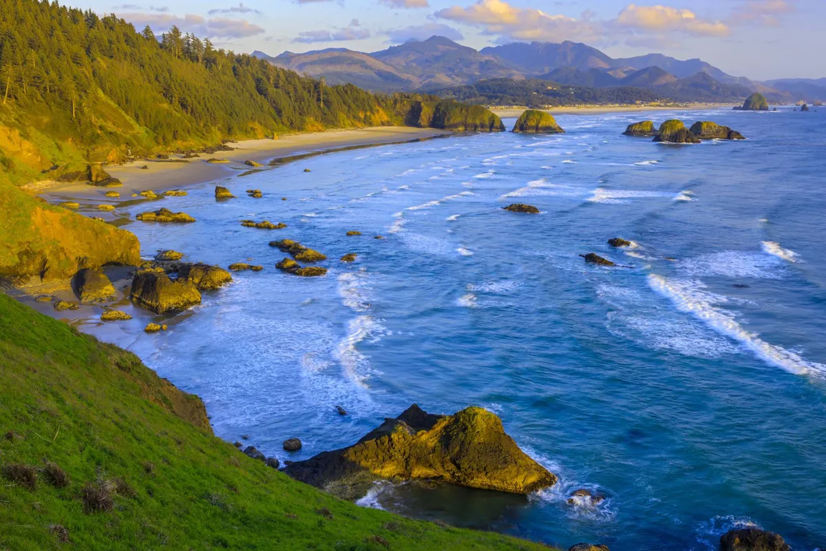 A panoramic view of a rocky coastline, with gentle waves rolling in and distant mountains framing the sunset sky. The scene is tranquil and picturesque, perfect for photography or simply soaking in the beauty of the Oregon coast.