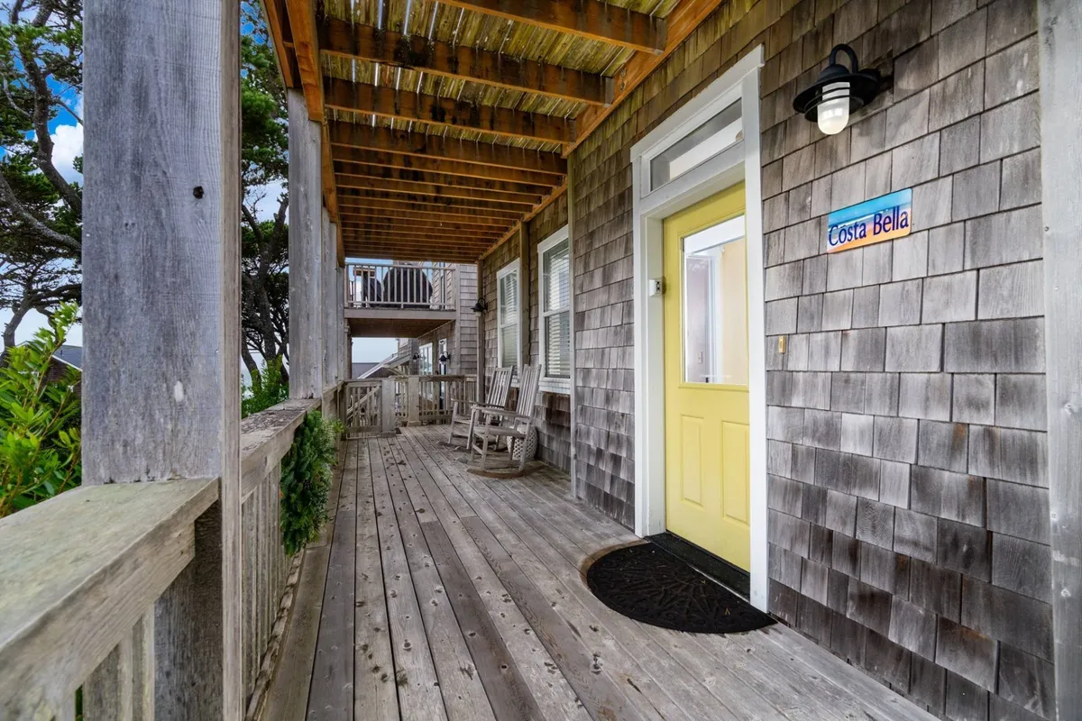 A cozy wooden deck with rocking chairs and a bright yellow door greets visitors at a cottage entrance. A coastal sign and lush greenery create a cheerful, inviting atmosphere perfect for family relaxation and neighborhood connection.