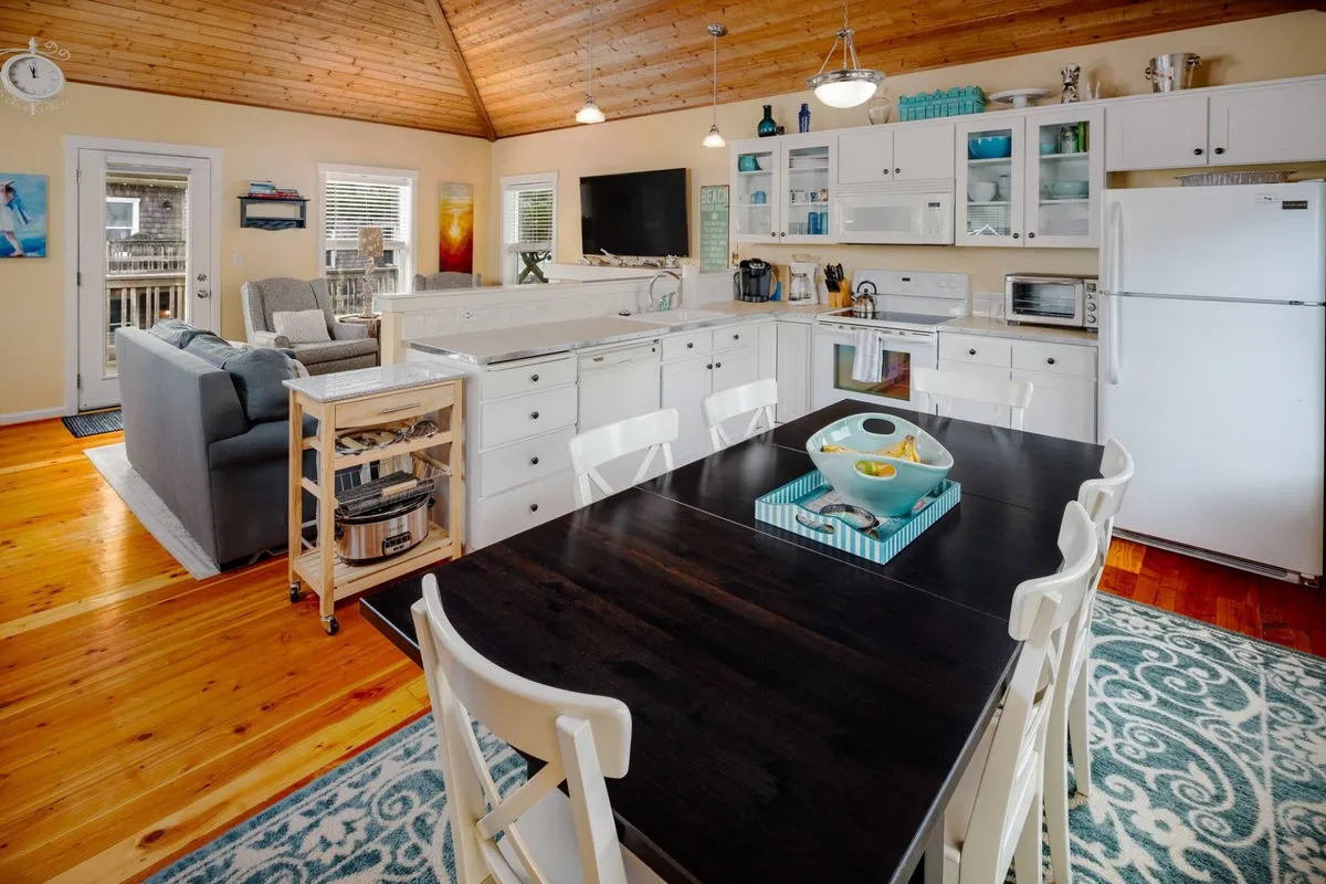 An open-plan space with a large dining table, a kitchen island, and a cozy living area with comfortable seating. Light pours in through windows, creating a welcoming environment that invites both relaxation and socializing.