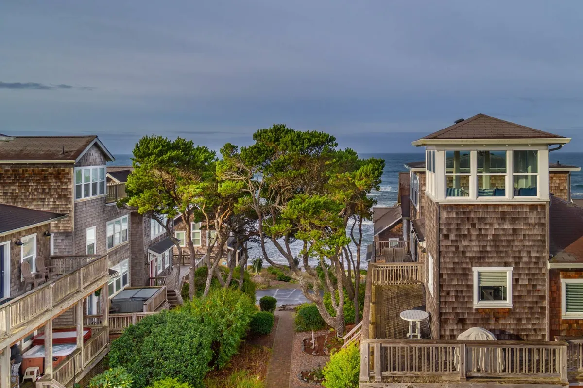 The image showcases the exterior of Costa Bella, with expansive decked patios overlooking the shimmering Pacific Ocean. A tranquil pathway winds through lush gardens and a picturesque tree line, inviting guests straight to the sandy beach below. The serene setting is perfect for sunset watching, relaxing to the sound of waves, or gathering around the fire pit with loved ones.