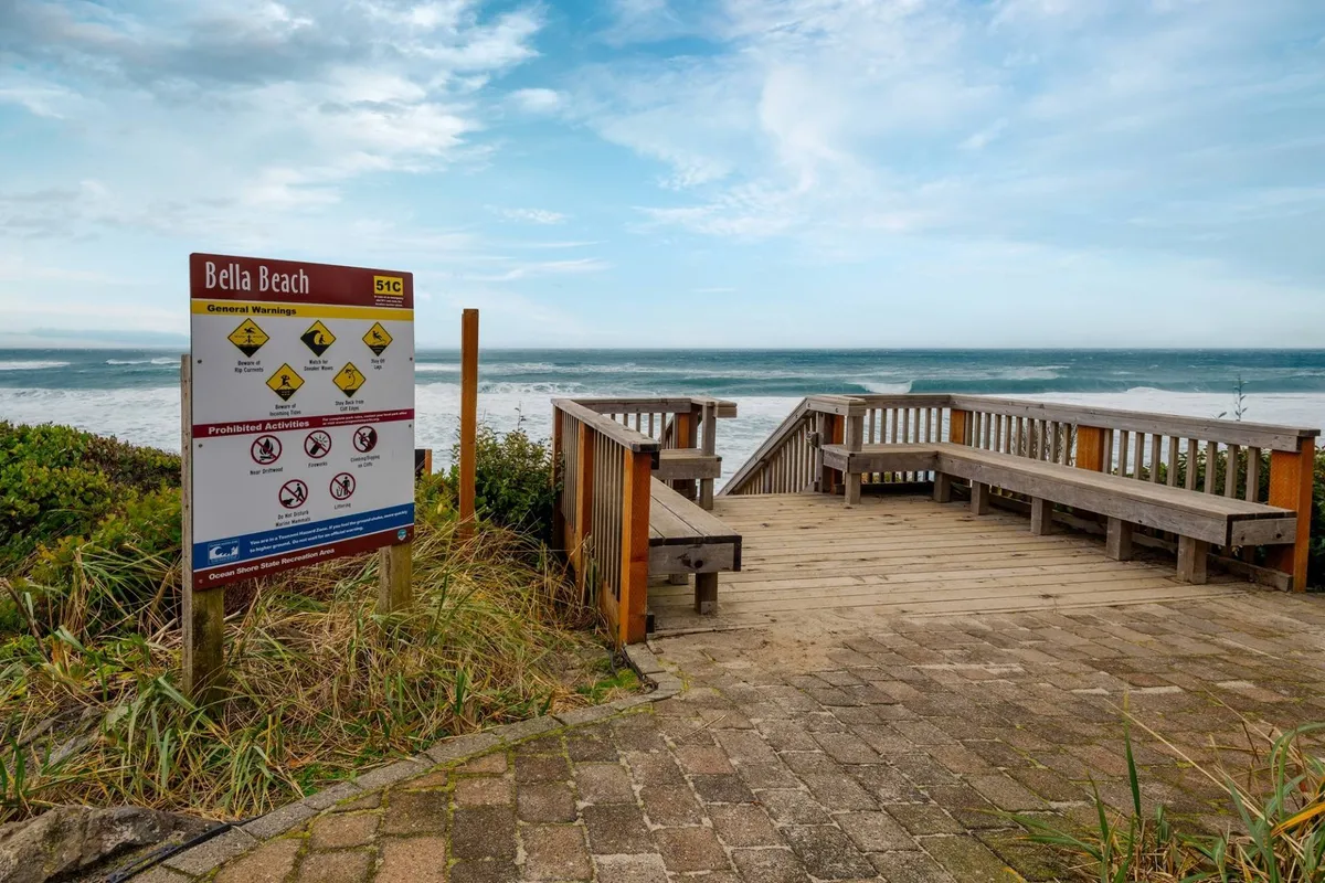 A wooden deck overlooks a sweeping ocean vista, with a 'beach' sign inviting guests toward the shoreline. The deck is bathed in soft, natural light, offering a tranquil setting for sunbathing, picnicking, or simply enjoying the sound of waves.