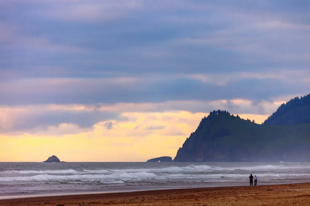 A couple walks hand-in-hand along a sandy beach as gentle ocean waves lap the shore. Distant cliffs provide a dramatic backdrop, and the soft glow of sunset casts a romantic light over the scene.