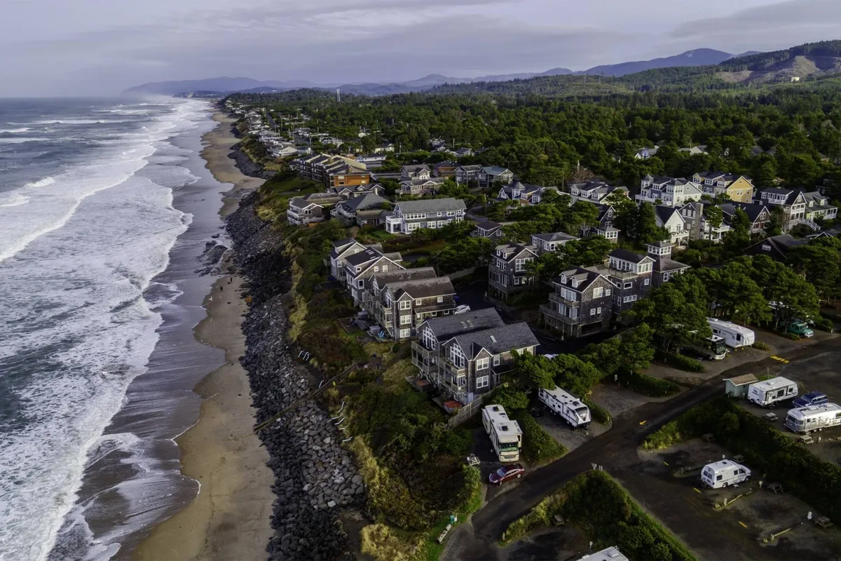 A wide view of the ocean waves rolling up to a sandy beach, with beachfront houses nestled along the shoreline and a lush forest as the backdrop. The scene evokes a sense of open community, natural beauty, and easy access to the beach.