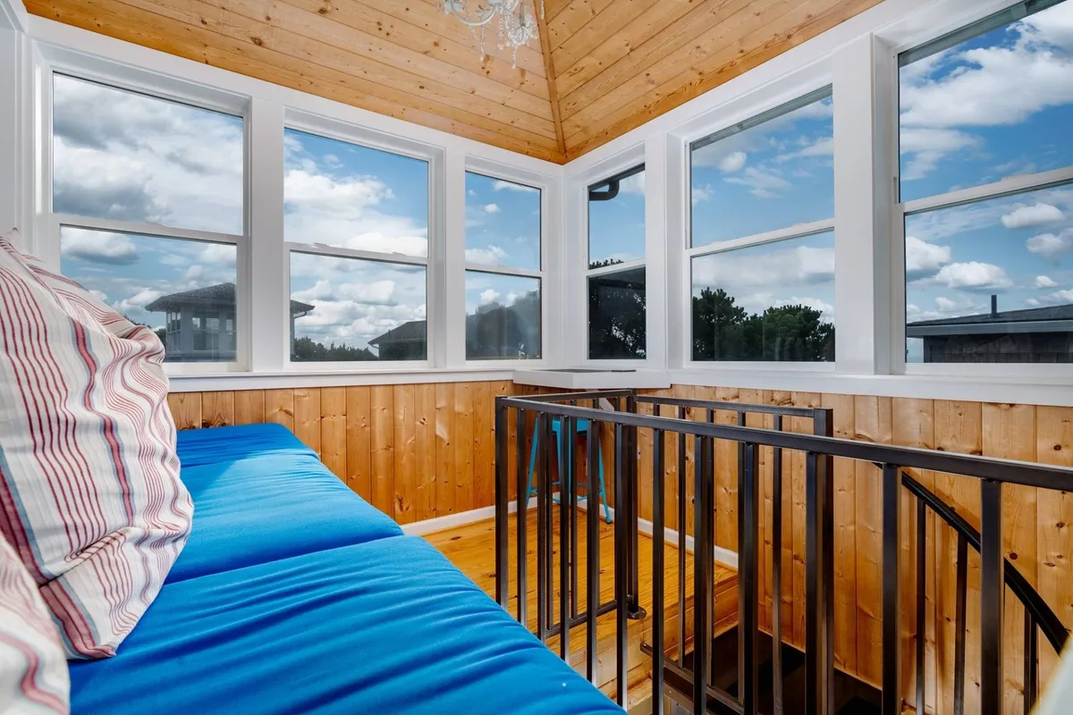 A calm, cozy interior with a blue couch set in front of large windows that let in abundant natural light, wood-paneled walls, and a modern railing. The view invites relaxation, reading, or quiet work, especially as the sun sets outside.
