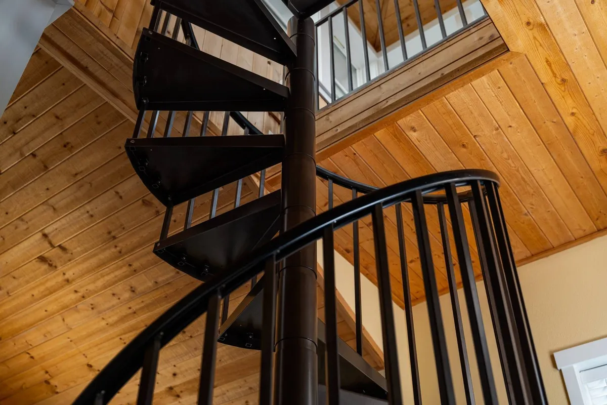 This image showcases the spiral staircase with its wooden ceiling and metal railing, creating an inviting ascent to the upper levels. The staircase is bathed in natural light, suggesting an airy, open feel that leads to a special sanctuary above.